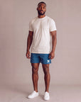 Male model wearing Mallard Blue MOVE Sport Apparel Agility 6 inch Training Shorts with a naturally untucked ivory tee in a neutral studio; original white thigh embellishment retained.