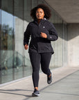 Woman running outdoors in the black MOVE Sport Apparel women’s AquaLight Packable Jacket and black leggings.