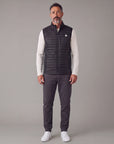 Man wearing the black MOVE Sport Apparel men’s down vest over an ivory long-sleeve top with charcoal trousers in a neutral studio.