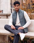 Man wearing the grey-green MOVE Sport Apparel Men’s down vest over an ivory long-sleeve top, seated on a stone bench beside autumn trees and architecture.