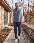 Man wearing the grey MOVE Sport Apparel Men’s down vest over a white long-sleeve top, walking beside stone architecture and autumn trees.