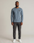 Full-body studio view of a man wearing the Blue Mirage Black Label long sleeve top with charcoal trousers and white trainers; original black square and white double-chevron chest embellishment retained.