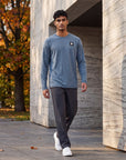 Man walking beside a stone building and autumn trees wearing the Blue Mirage Black Label long sleeve top with charcoal trousers and white trainers; original chest embellishment retained.