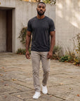 Man walking through a stone courtyard in the Asphalt MOVE Sport Apparel men’s Merino Short Sleeve with stone trousers and white trainers; original sleeve tab retained.