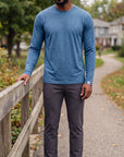 Man wearing the MOVE Sport Apparel Men’s Essential Long Sleeve in Blue Mirage with charcoal trousers beside a wooden railing on a leafy park path.