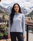 A woman wearing a MOVE sport apparel light blue essential quarter zip with a reflective logo on a mountain balcony.