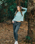 MOVE sport apparel woman wearing a light blue long sleeve crewneck sweatshirt and dark grey joggers in a forest.