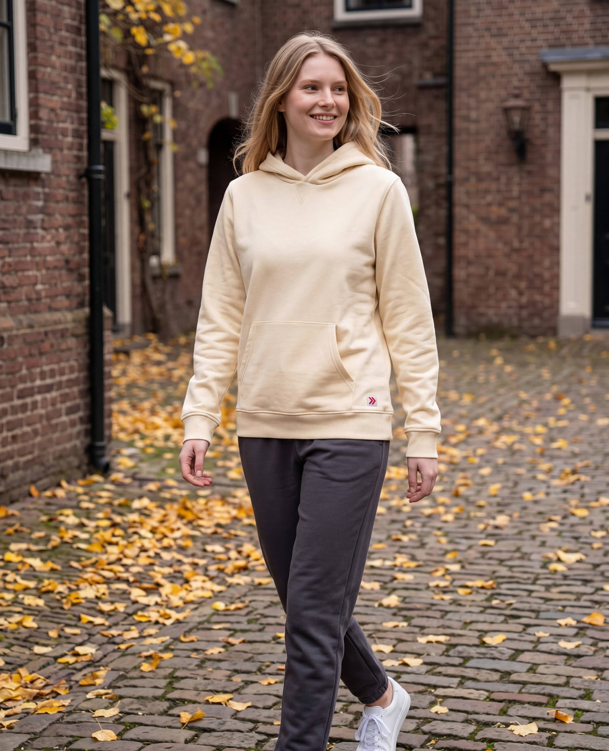 Woman walking through a brick courtyard in autumn wearing the MOVE Sport Apparel French Terry Hoodie in Natural.