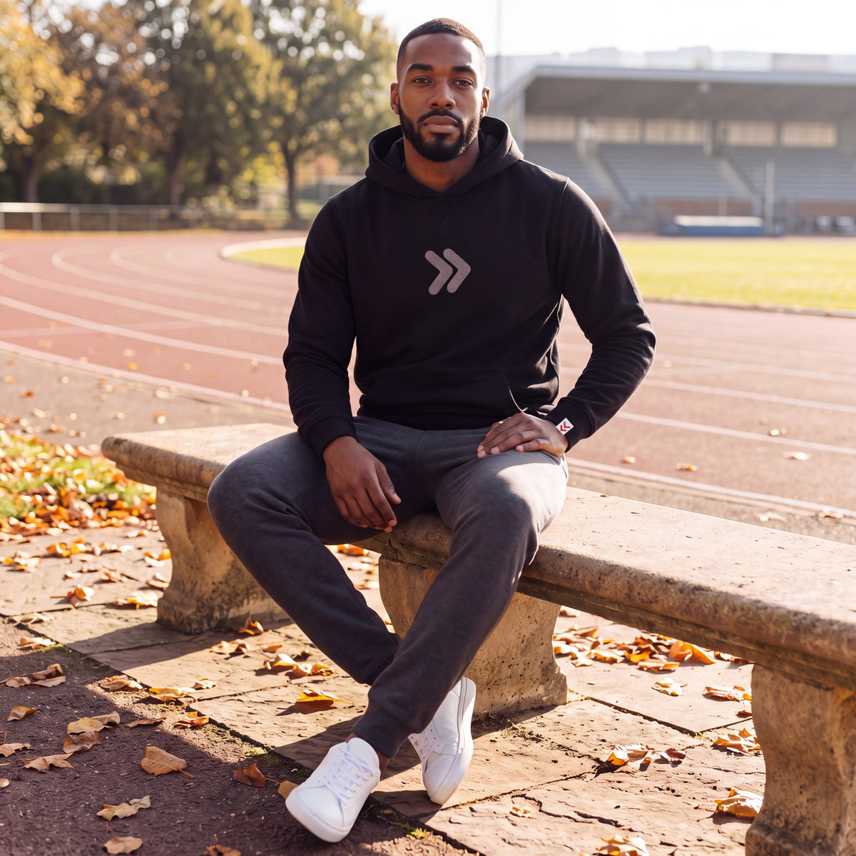 Man wearing the black MOVE Sport Apparel Logo Hoodie with grey chest chevrons and the original cuff tab, seated beside an outdoor running track in autumn.