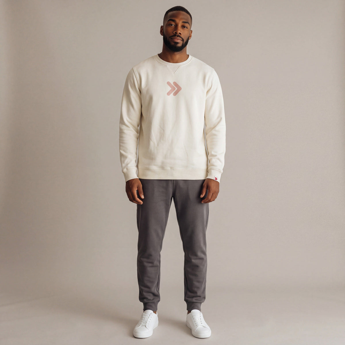 Male model wearing the cream MOVE Sport Apparel Logo Sweatshirt with charcoal joggers in a neutral studio; original blush-pink centered chest embellishment and red-and-white cuff tab retained.