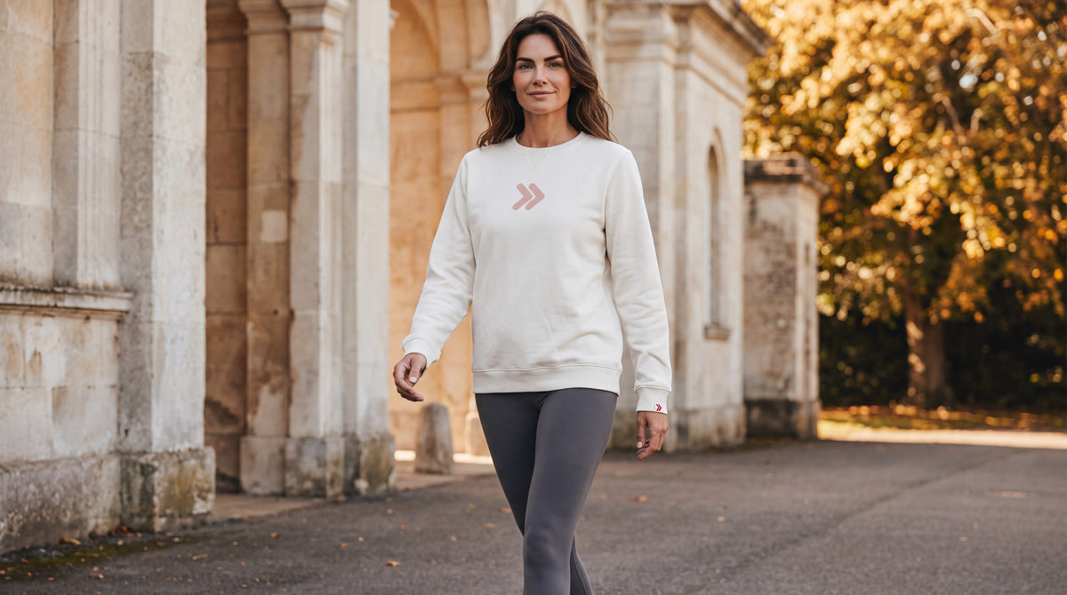 Woman wearing the cream MOVE Sport Apparel Logo Sweatshirt with charcoal leggings beside stone architecture and autumn trees.