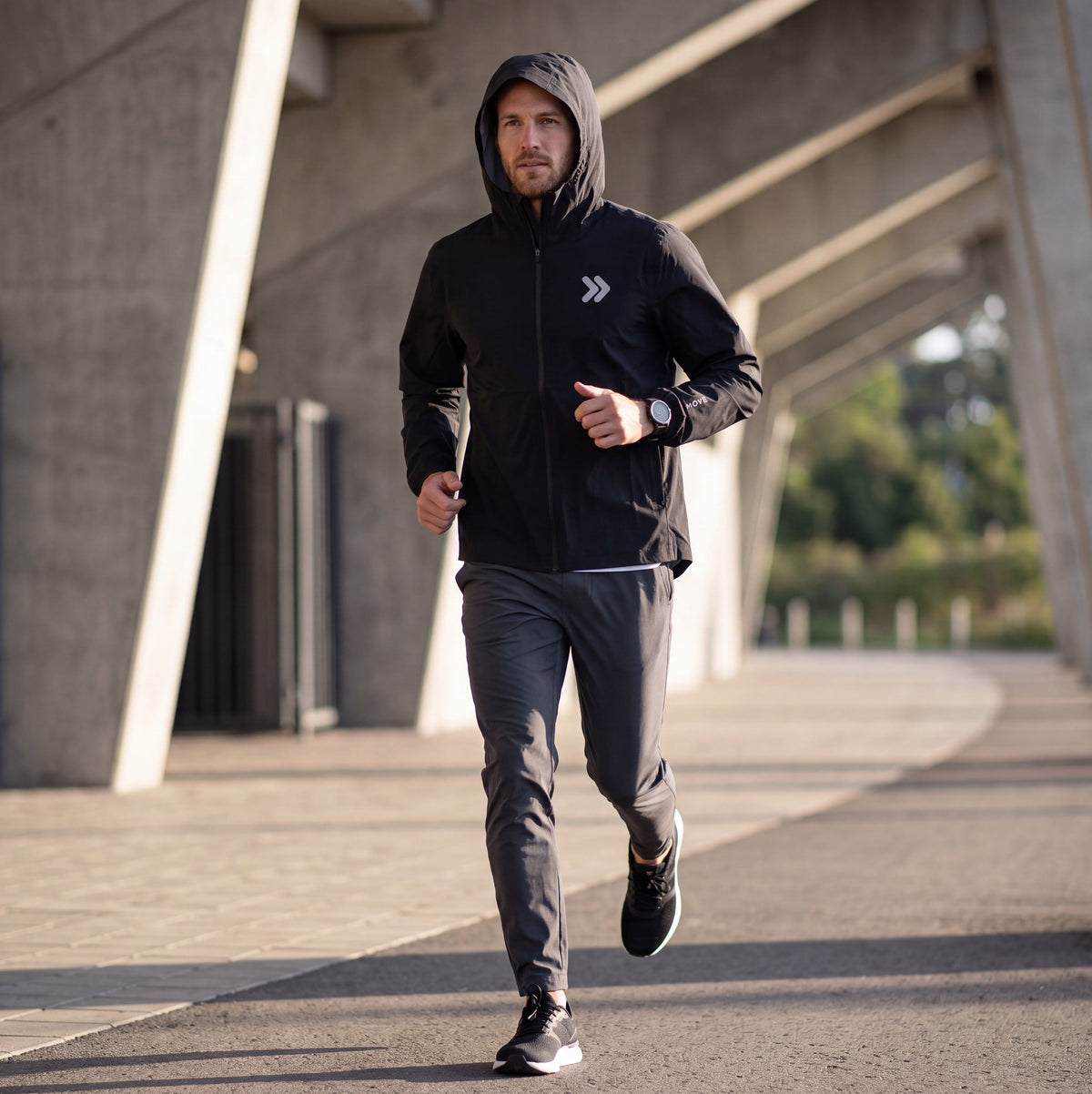 Man running on an outdoor concrete path in the black MOVE Sport Apparel men’s AquaLight hooded jacket.