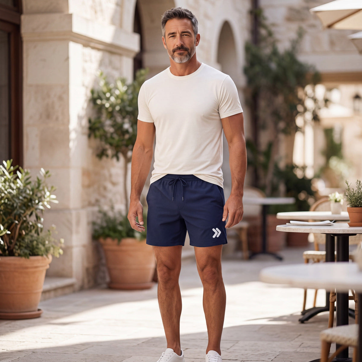 MOVE Sport Apparel Men’s Core Athletic Training Shorts in Naval Academy navy, worn by an adult man with a lightweight ivory athletic tee partially tucked at the front, in a sunlit outdoor cafe setting.