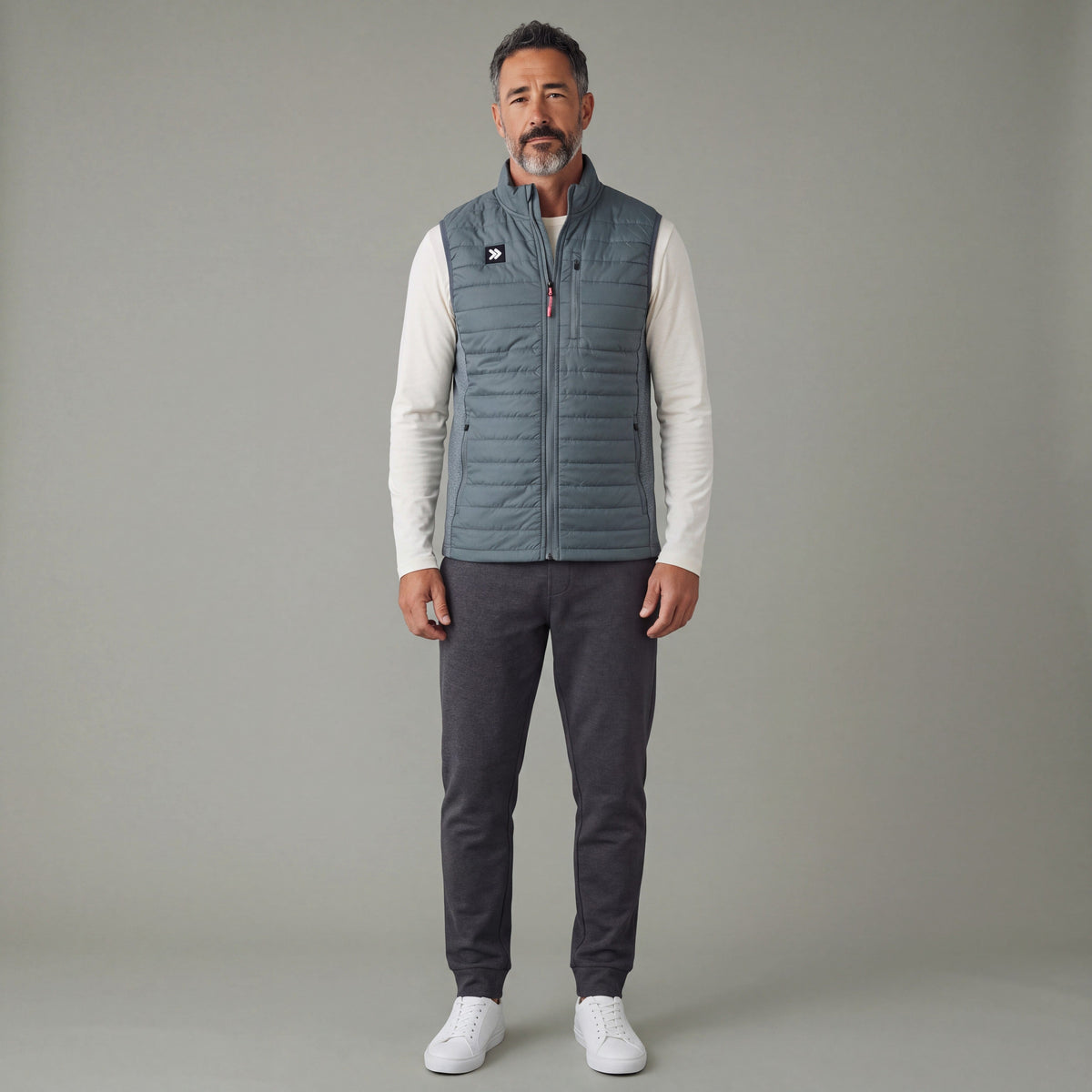 Male model wearing the grey-green MOVE Sport Apparel Men’s down vest over an ivory long-sleeve tee with charcoal joggers in a neutral studio; original chest patch, quilting and zipper details retained.