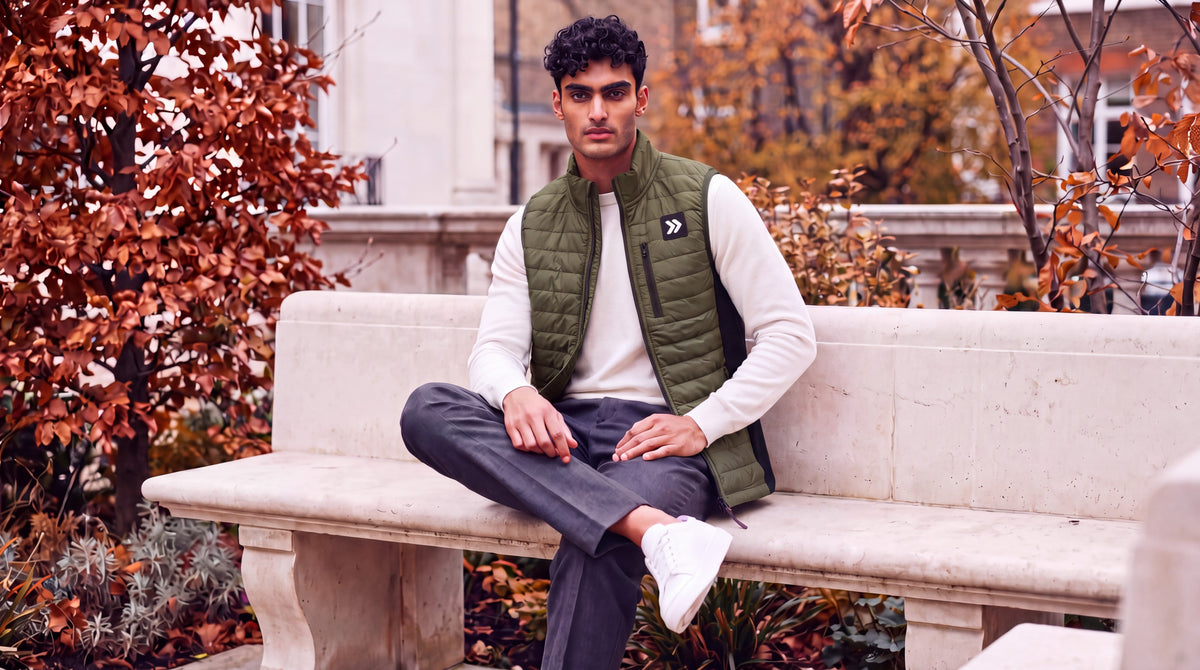 Man wearing the olive Winter Moss MOVE Sport Apparel men’s down vest over an ivory long-sleeve top, seated on a stone bench beside autumn trees.