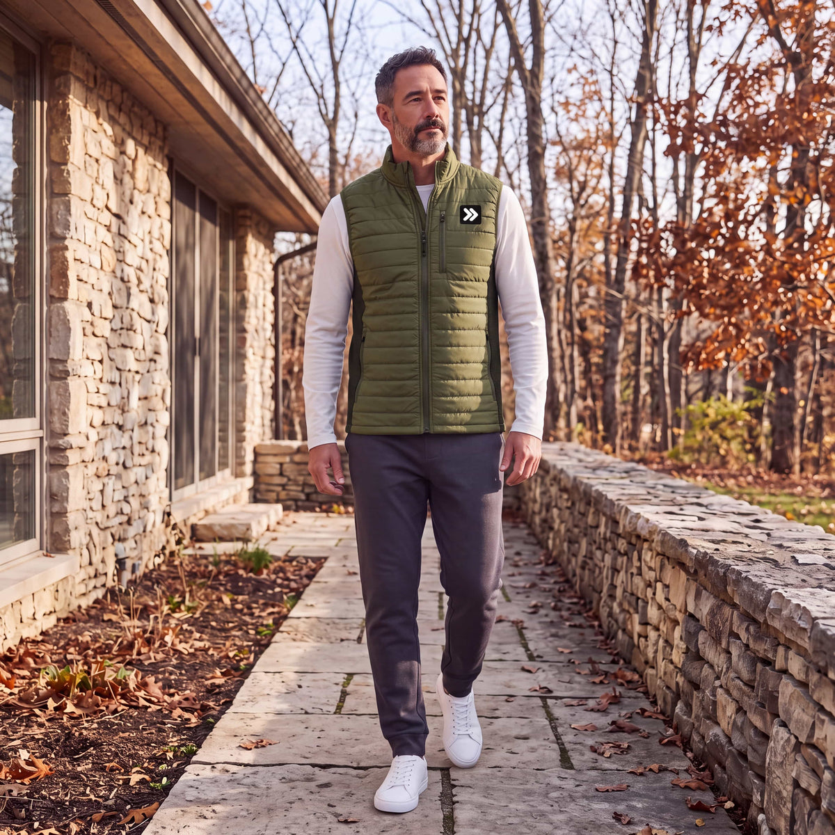 Man wearing the olive Winter Moss MOVE Sport Apparel men’s down vest over an ivory long-sleeve top, walking beside stone architecture and autumn trees.