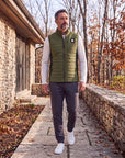Man wearing the olive Winter Moss MOVE Sport Apparel men’s down vest over an ivory long-sleeve top, walking beside stone architecture and autumn trees.