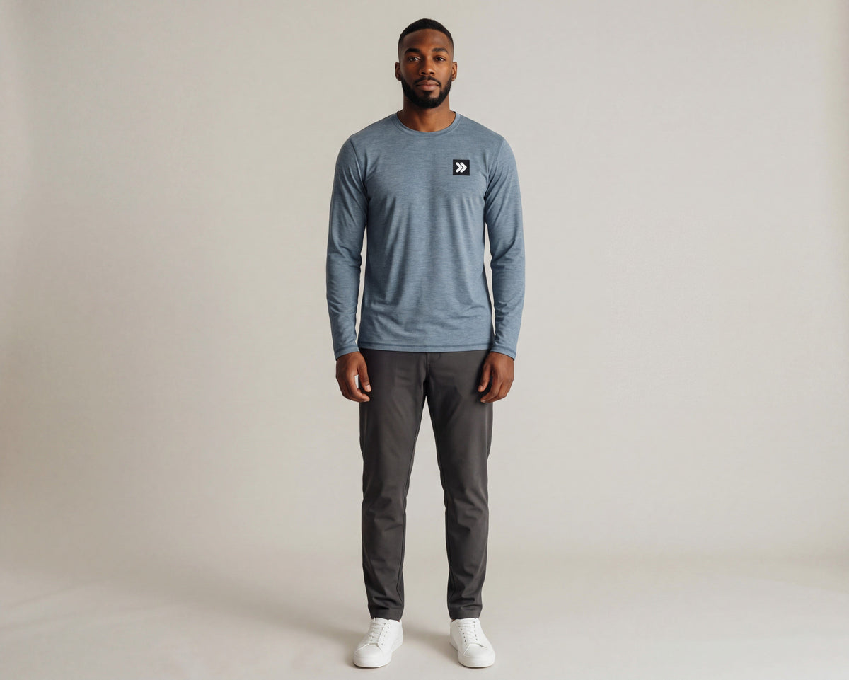 Full-body studio view of a man wearing the Blue Mirage Black Label long sleeve top with charcoal trousers and white trainers; original black square and white double-chevron chest embellishment retained.
