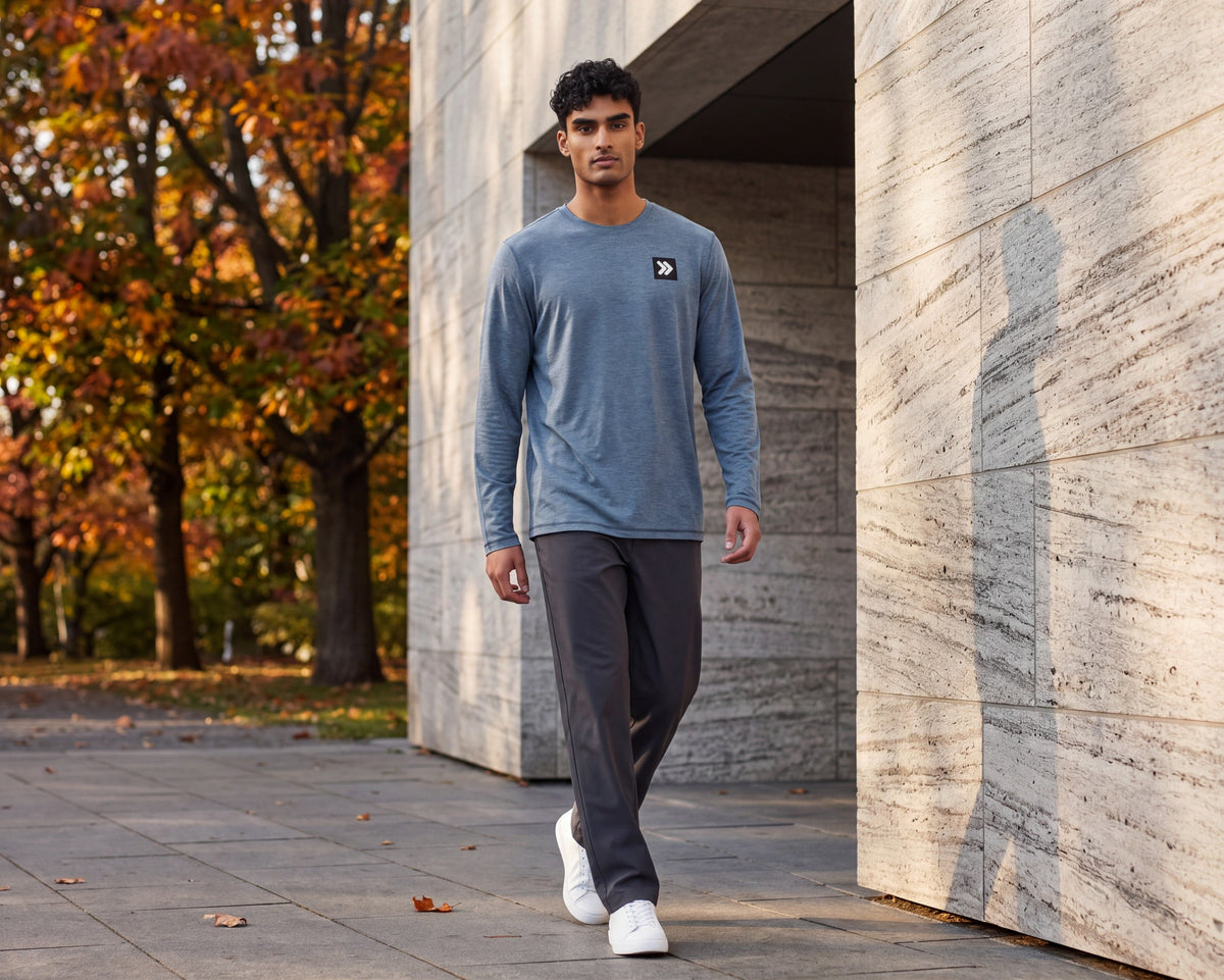 Man walking beside a stone building and autumn trees wearing the Blue Mirage Black Label long sleeve top with charcoal trousers and white trainers; original chest embellishment retained.