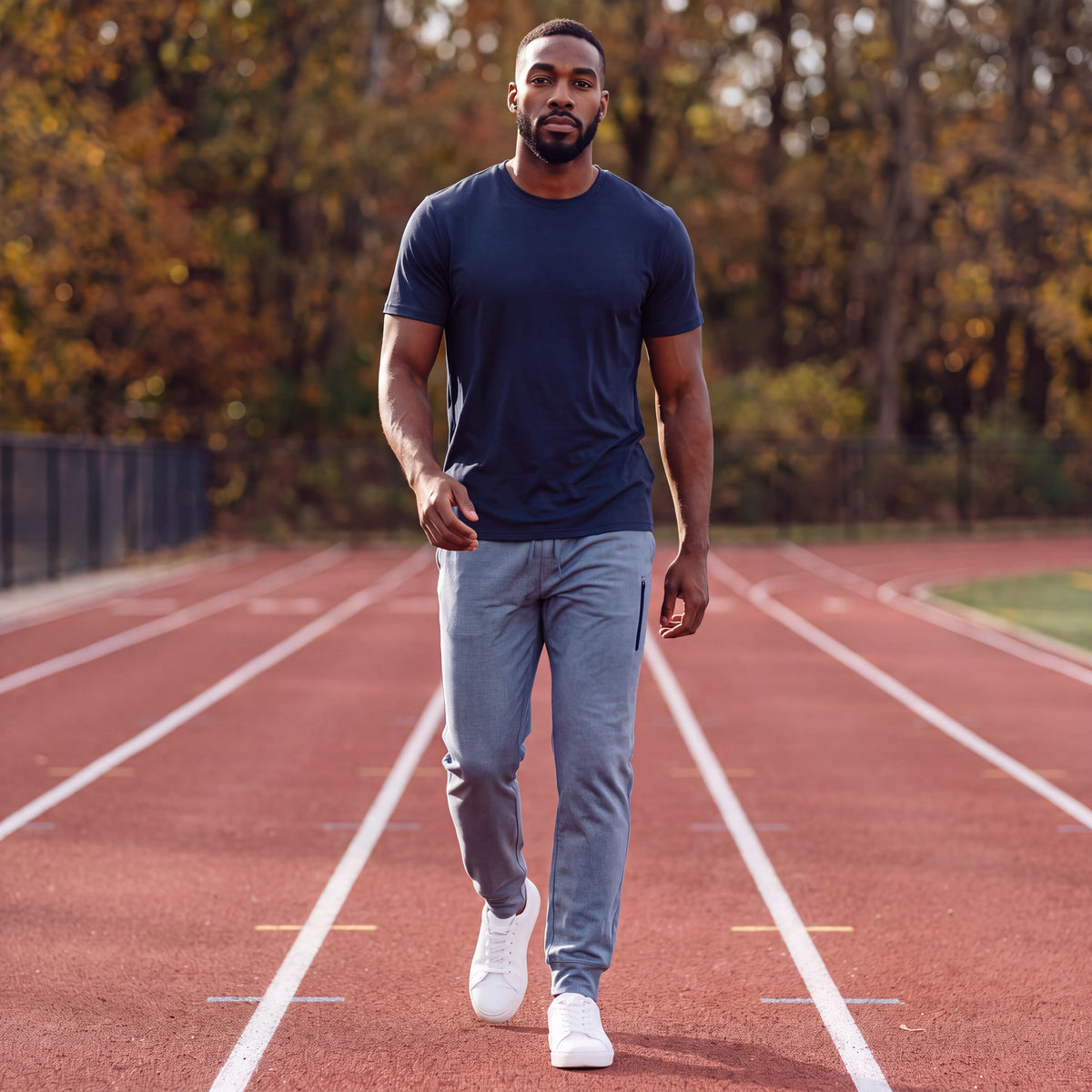 Man warming up on an outdoor track in autumn wearing Blue Mirage MOVE Sport Apparel men’s Essential Jogger with a plain untucked navy tee and white trainers.