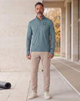Man wearing the Heather North Atlantic MOVE Sport Apparel Essential Quarter Zip Pro with sand trousers and off-white sneakers at a limestone athletic recovery terrace beside an autumn running track.