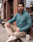 Man wearing the Heather North Atlantic MOVE Sport Apparel Essential Quarter Zip Pro with sand trousers, seated outside a coffee shop.