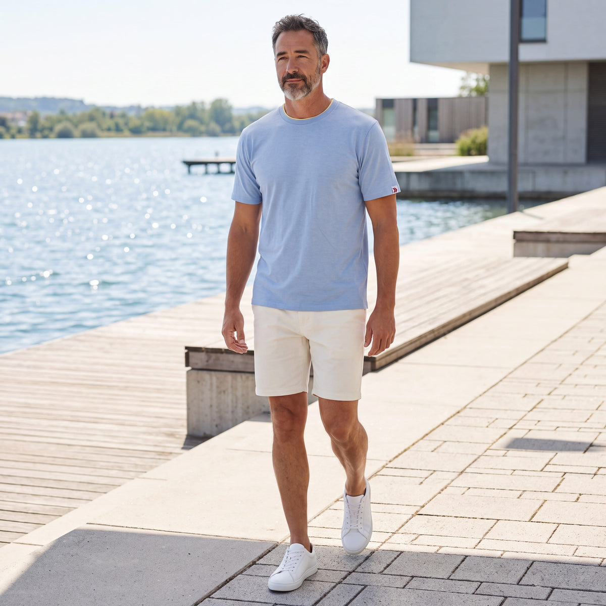 Man wearing MOVE Sport Apparel men’s Essential Short Sleeve in Heather Chambray Blue with cream shorts while walking along a sunlit waterfront promenade.