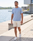 Man wearing MOVE Sport Apparel men’s Essential Short Sleeve in Heather Chambray Blue with cream shorts while walking along a sunlit waterfront promenade.