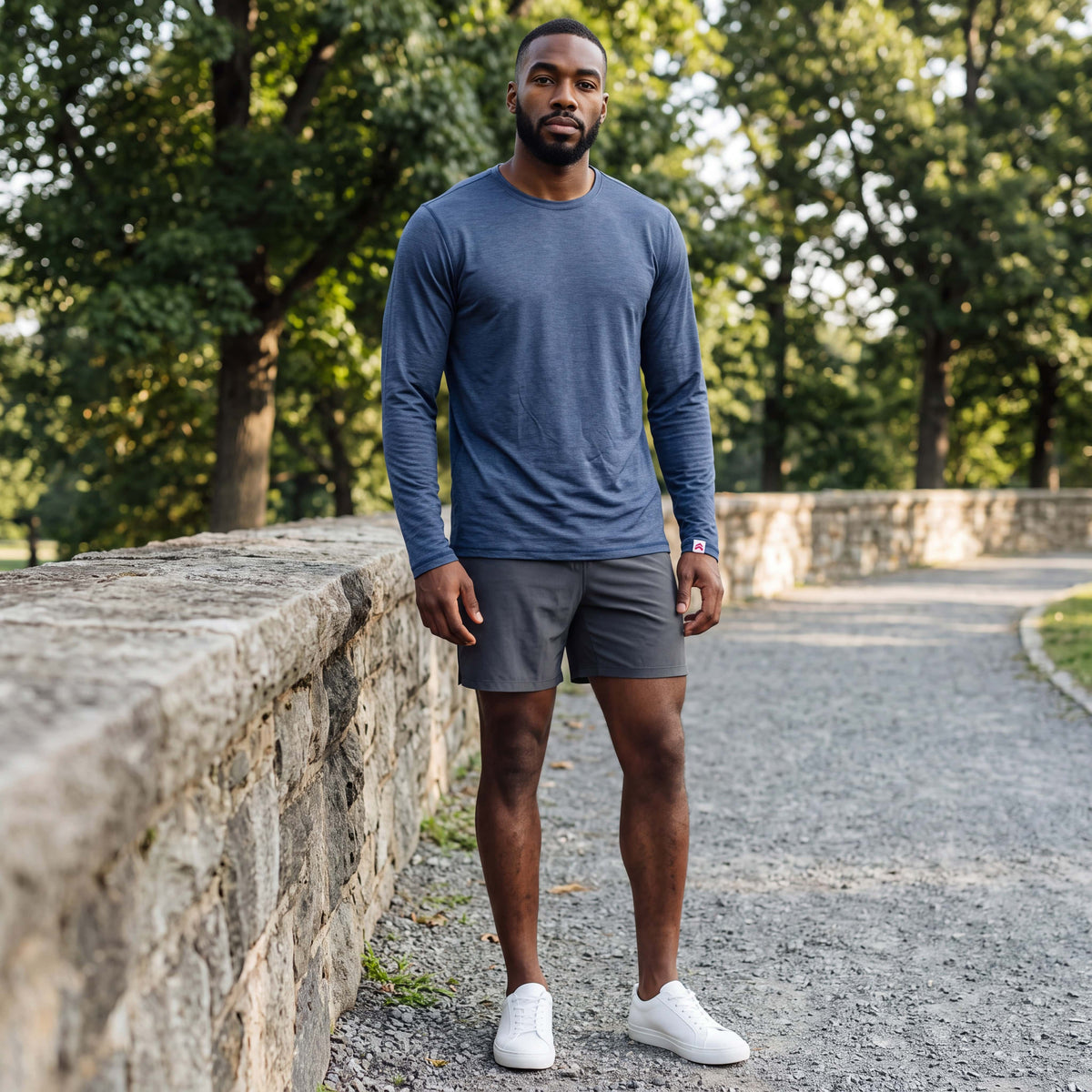 Man wearing MOVE Sport Apparel men’s LuxDry long sleeve in Naval academy with charcoal shorts beside a stone wall on a wooded park path.