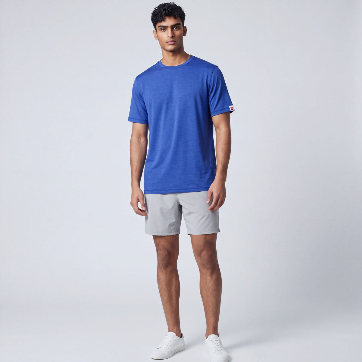 Full-body studio view of a man wearing the Surf the Web blue LuxDry Training T-Shirt with light grey shorts and white trainers; original sleeve tab retained.