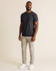 Full-body studio view of a man wearing the Asphalt MOVE Sport Apparel men’s Merino Short Sleeve with stone trousers and white trainers; original red-double-chevron sleeve tab retained.