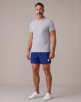 Male model wearing Beacon Blue MOVE Sport Apparel men’s Performance 5 Inch Running Shorts with an untucked light-grey athletic T-shirt in a neutral studio; original white thigh embellishment retained.