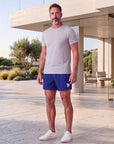 Man wearing Beacon Blue MOVE Sport Apparel Performance 5 Inch Running Shorts with an untucked light-grey tee and white trainers on a sunlit limestone terrace beside coastal architecture; short hem and white thigh embellishment visible.