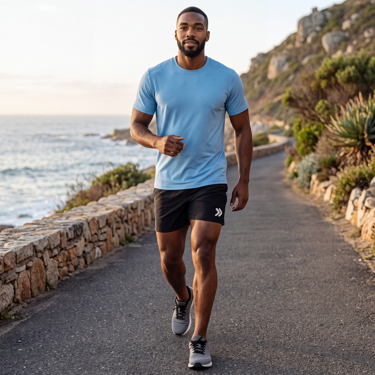 Man warming up beside a coastal running path in black MOVE Sport Apparel men’s Performance 5 Inch Running Shorts with an untucked sky-blue tee; original white thigh embellishment retained.