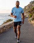 Man warming up beside a coastal running path in black MOVE Sport Apparel men’s Performance 5 Inch Running Shorts with an untucked sky-blue tee; original white thigh embellishment retained.