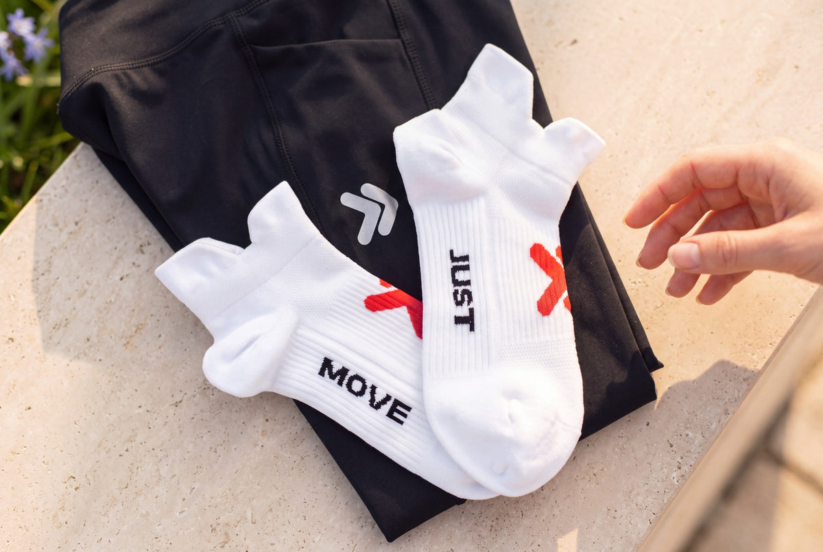 White MOVE Sport Apparel Performance Lightweight Ankle Socks with red embellishments and black JUST MOVE lettering, arranged over a black garment on sunlit limestone beside greenery.