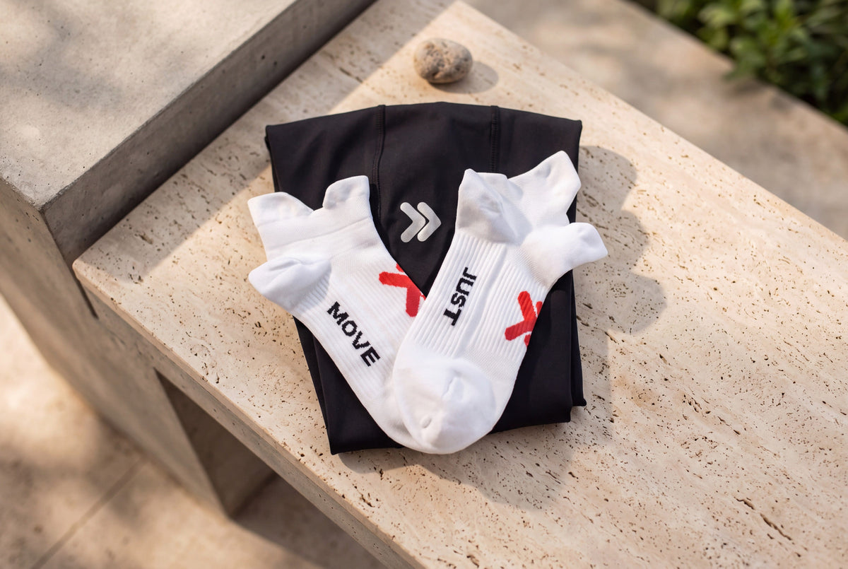 White MOVE Sport Apparel Performance Midweight Double Tab Socks with raised ankle tabs, red embellishments and black JUST MOVE lettering, arranged over a black garment on a sunlit travertine bench.