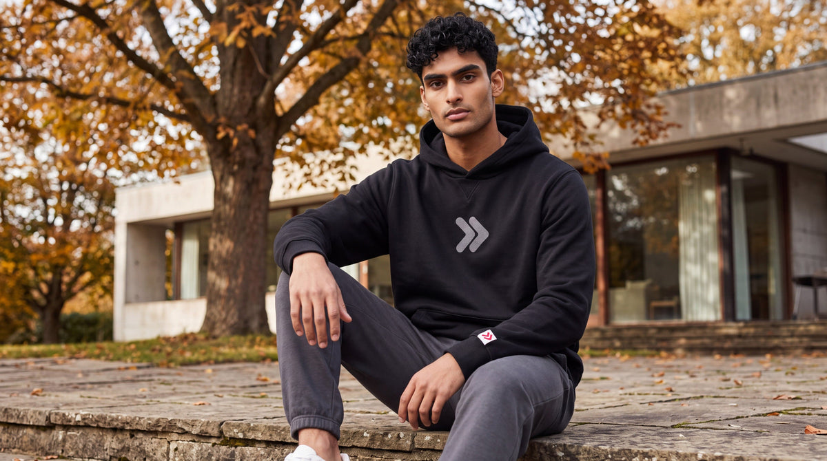 Man seated on a stone terrace wearing the black MOVE Sport Apparel Logo Hoodie and charcoal joggers, with autumn trees and contemporary architecture behind him.