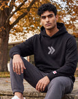 Man seated on a stone terrace wearing the black MOVE Sport Apparel Logo Hoodie and charcoal joggers, with autumn trees and contemporary architecture behind him.