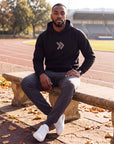 Man wearing the black MOVE Sport Apparel Logo Hoodie with grey chest chevrons and the original cuff tab, seated beside an outdoor running track in autumn.