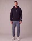 Man wearing the black MOVE Sport Apparel Logo Hoodie with slate-grey joggers in a neutral studio, showing the small centered grey rounded double chevrons and red-and-white tab on the left sleeve cuff.