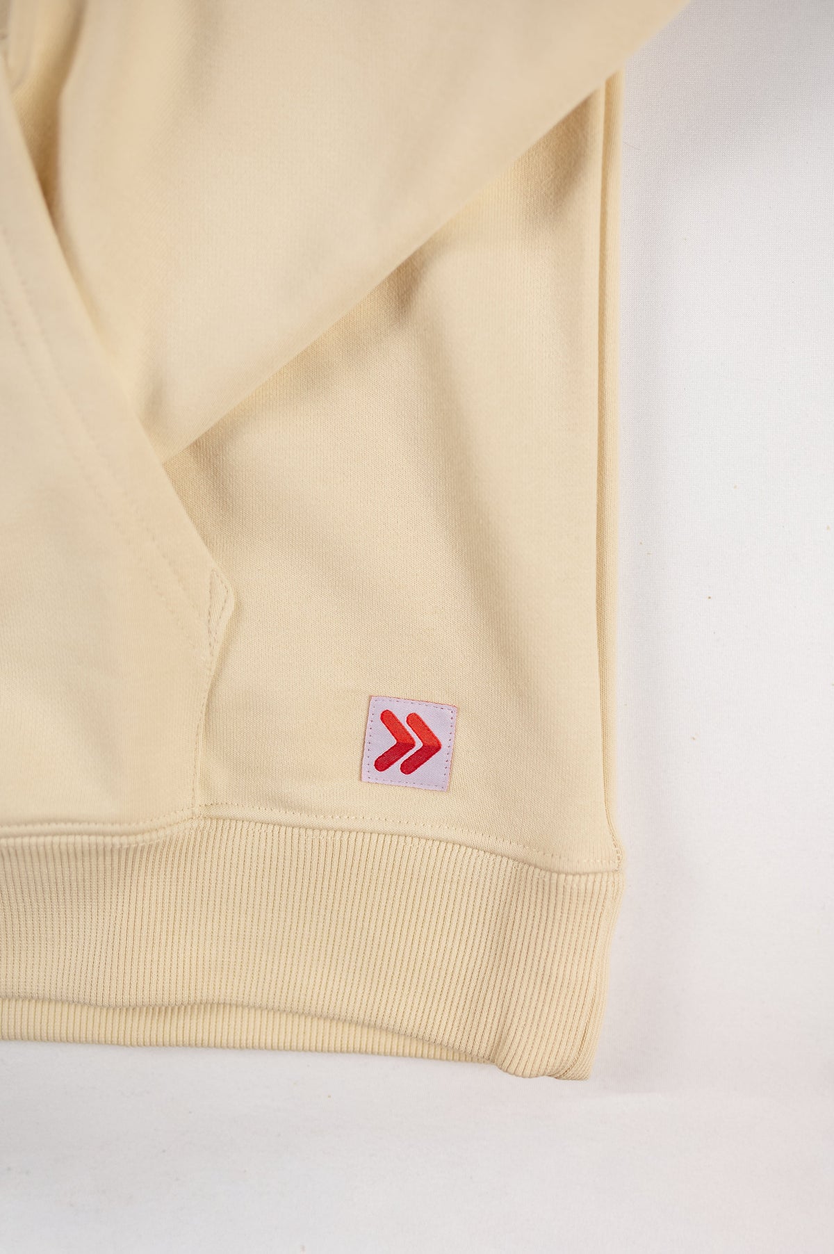 Close view of the original red double chevrons on the white hem label of the MOVE Sport Apparel French Terry Hoodie in Natural.