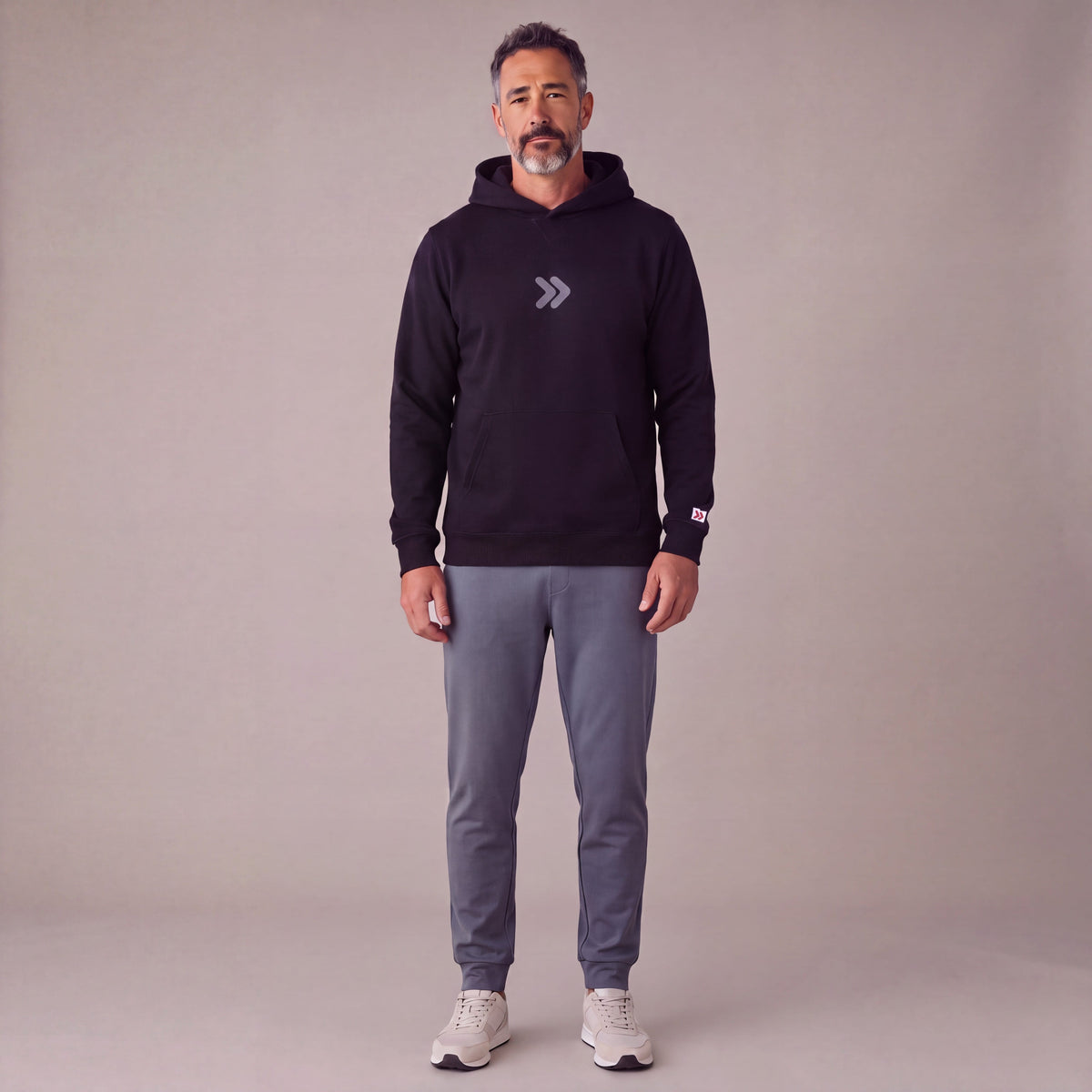 Man wearing the black MOVE Sport Apparel Logo Hoodie with slate-grey joggers in a neutral studio, showing the small centered grey rounded double chevrons and red-and-white tab on the left sleeve cuff.