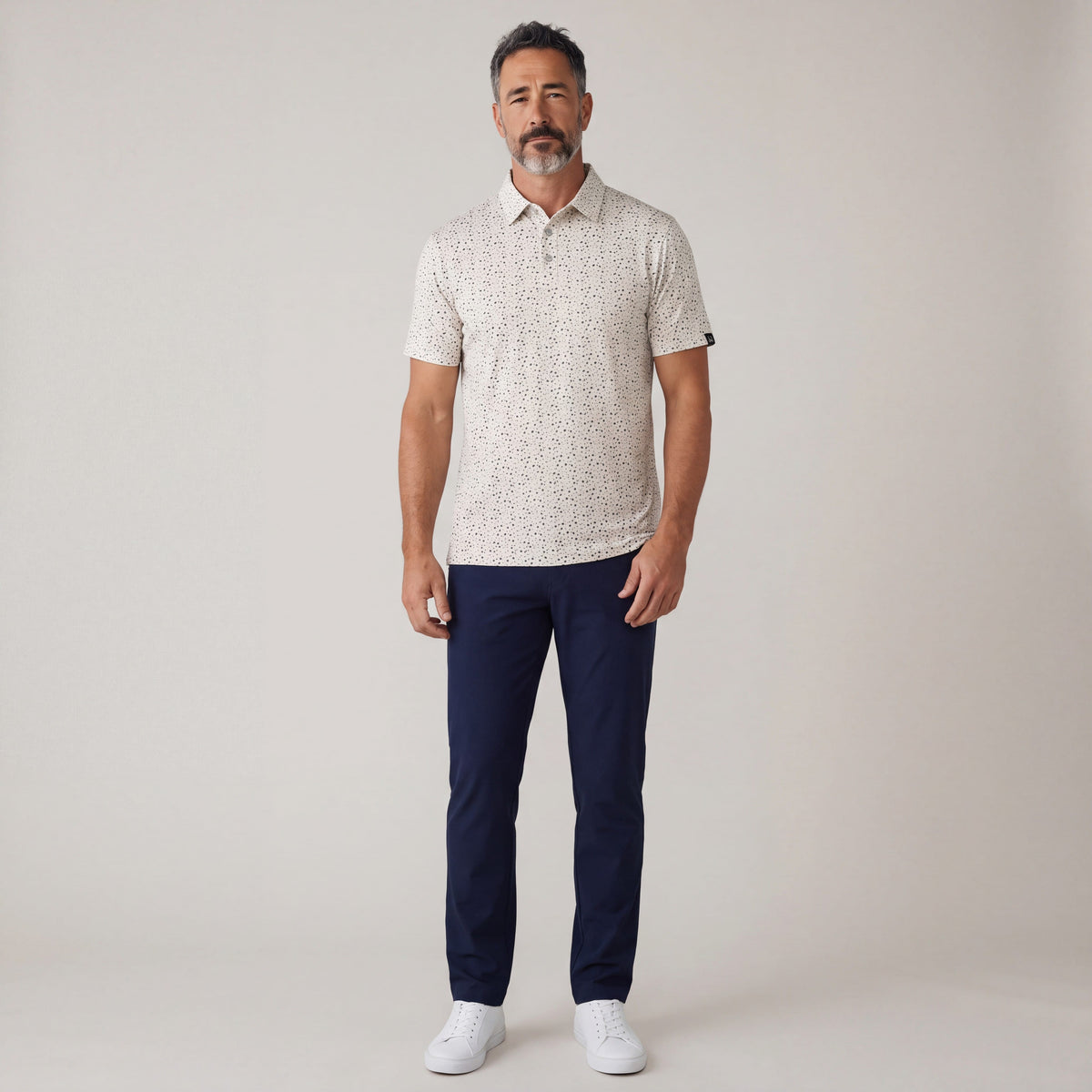 MOVE Sport Apparel Men’s Clubhouse Golf Polo in cream On Par black-fleck print, worn untucked with navy trousers by an adult man in a neutral full-body studio view.