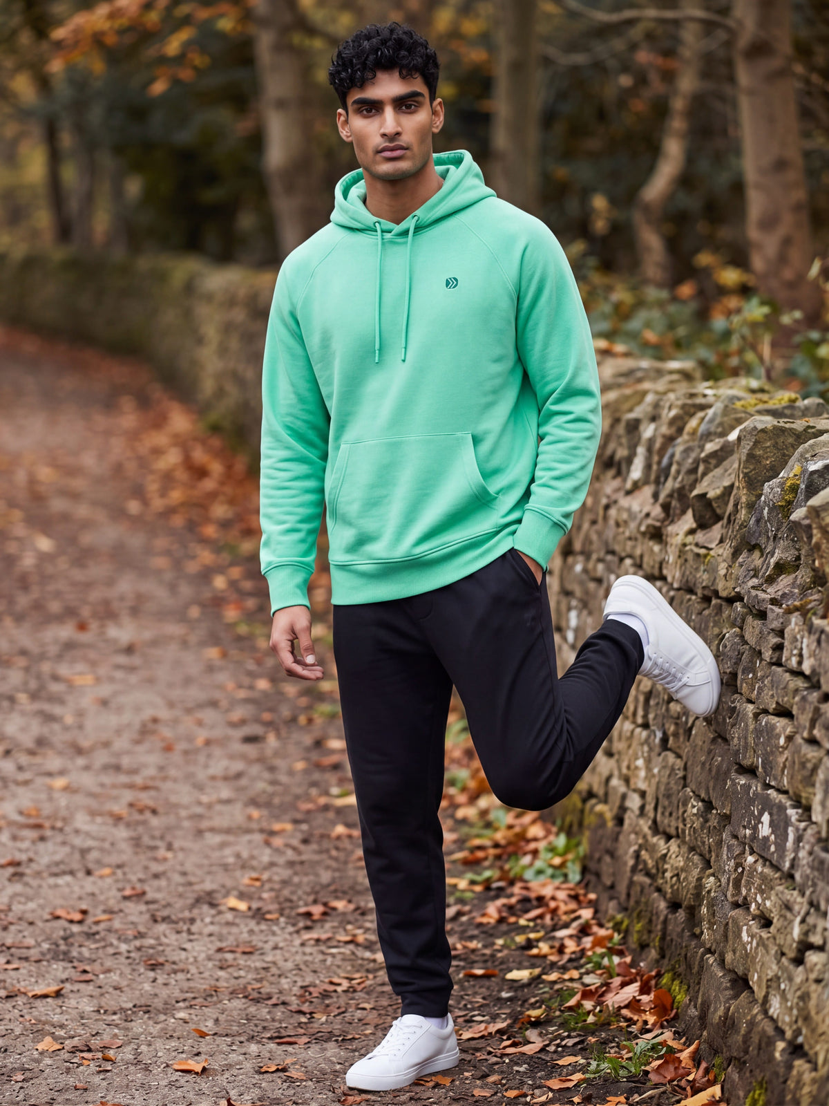 MOVE Sport Apparel Men’s Essential Pullover Hoodie in Blarney mint green, worn by an adult man stretching beside a stone wall on an autumn trail.