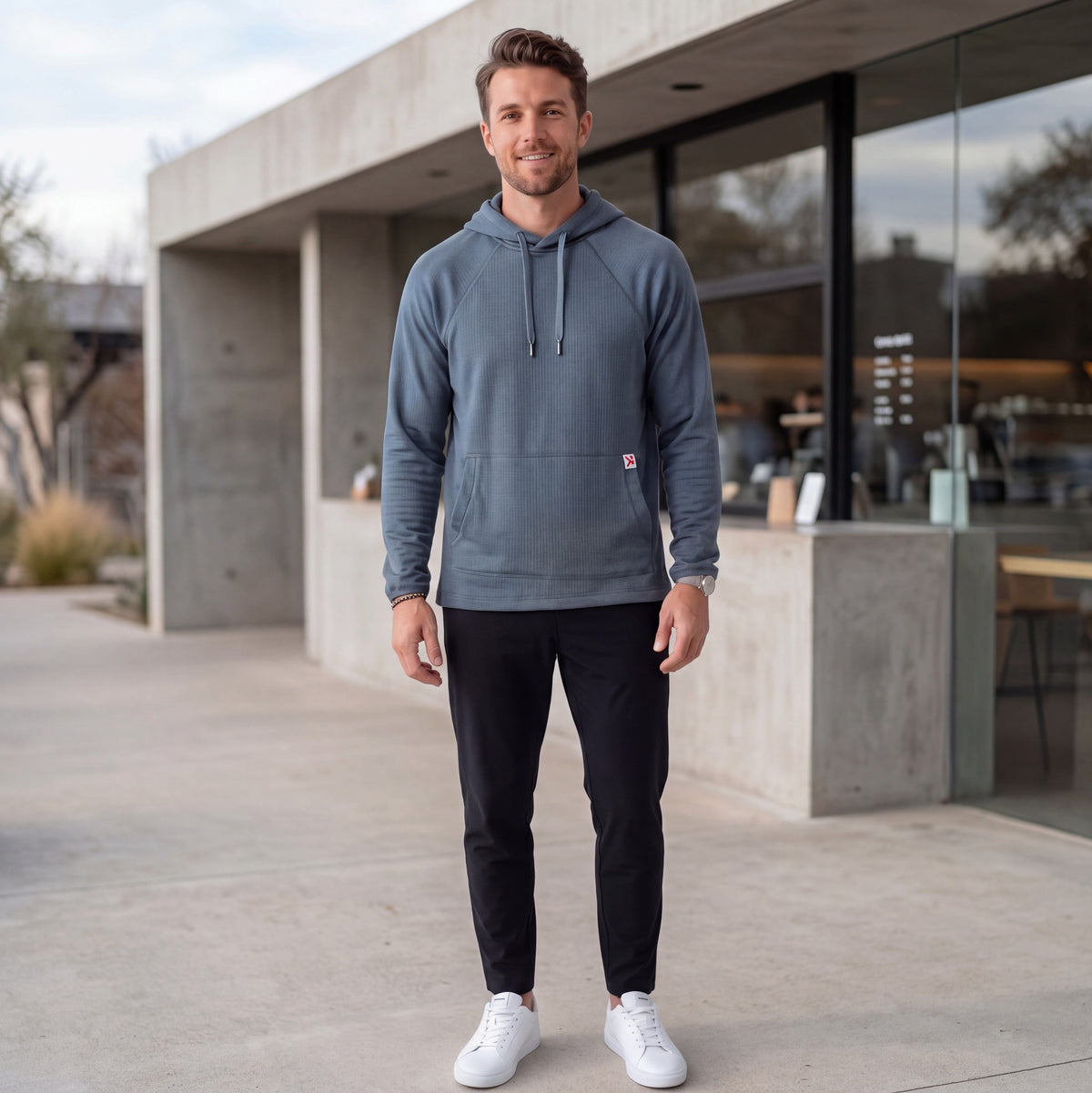 Athletic male model wearing the MOVE Sport Apparel men’s waffle hoodie in Blue Heather Ashes outside a modern cafe, with black training pants and white athletic shoes.