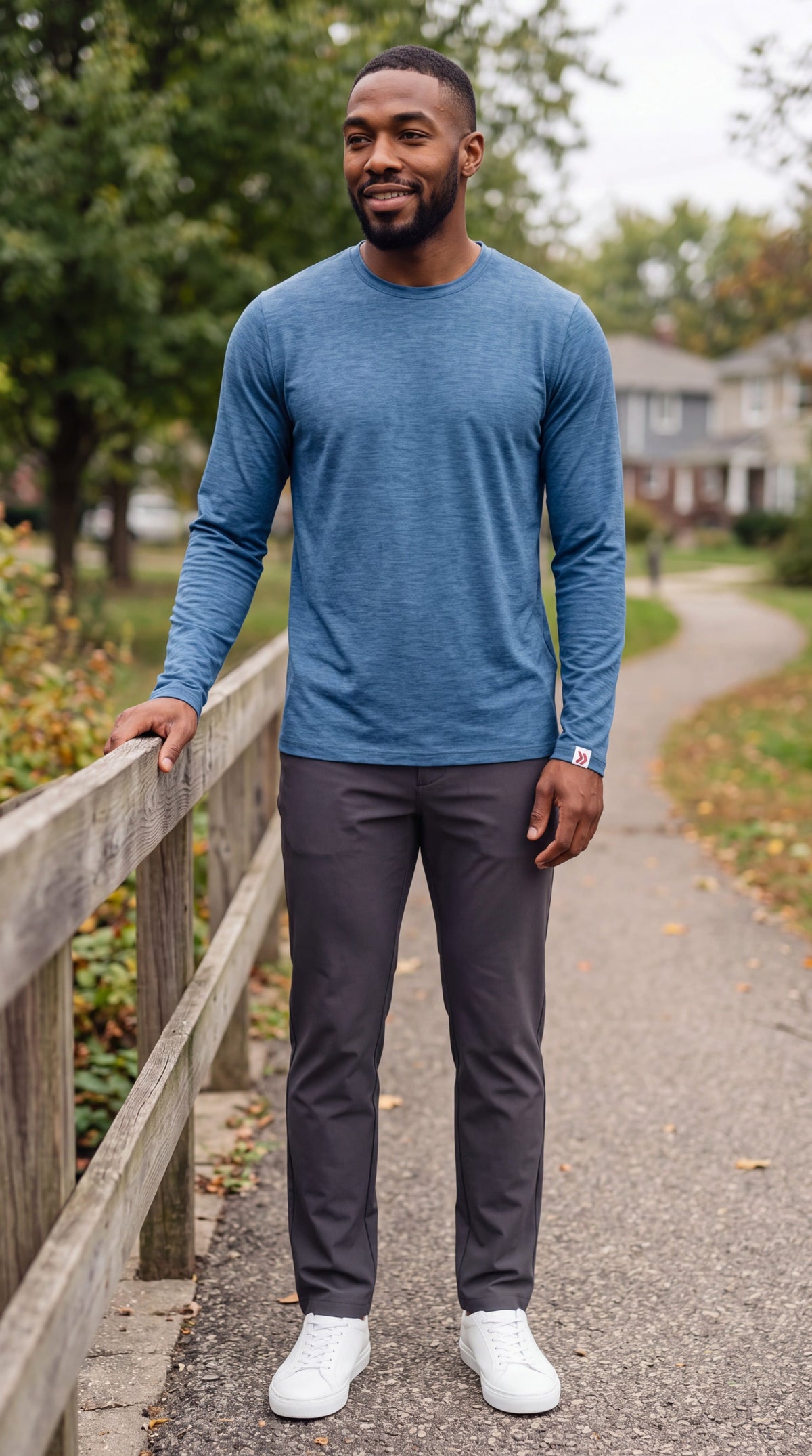 Male model wearing the Blue Mirage MOVE Sport Apparel Essential Long Sleeve crewneck and charcoal trousers beside a wooden railing on a leafy park path.