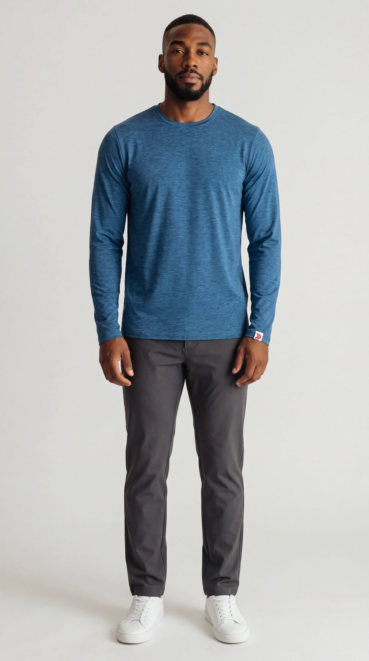 Full-body male model wearing the Blue Mirage MOVE Sport Apparel Essential Long Sleeve crewneck with charcoal trousers and white sneakers in a neutral studio.