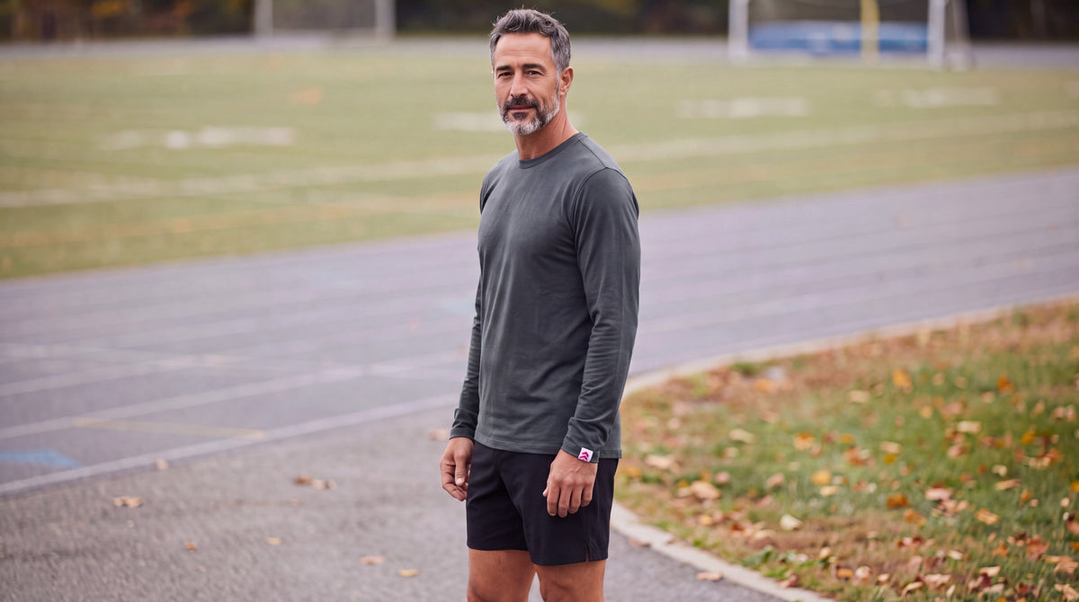 Man wearing the MOVE Sport Apparel Men’s Essential Long Sleeve in Heather Darkest Spruce with black training shorts beside an outdoor running track in autumn.