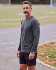 Man wearing the MOVE Sport Apparel Men’s Essential Long Sleeve in Heather Darkest Spruce with black training shorts beside an outdoor running track in autumn.